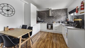 A modern kitchen with white cabinets, a wooden dining table, black chairs, a large clock, and a grey marble backsplash. - Comfort behind Crouch In CBD, newly renovated, perfect for the family & pets (Mount Gambier)