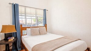 A bedroom with a bed, a bedside table with a lamp, and curtains. - Mango Paradise · Steps from the Beach & Fast Wi-Fi (Jacó)