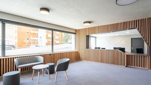 Zona de recepción con un diseño moderno, que incluye un mostrador de recepción, un área de descanso con un banco y sillas, y ventanas amplias con vistas a la calle.