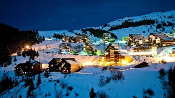 Un pueblo montañoso cubierto de nieve en la noche, con edificios iluminados y una pista de esquí al fondo.