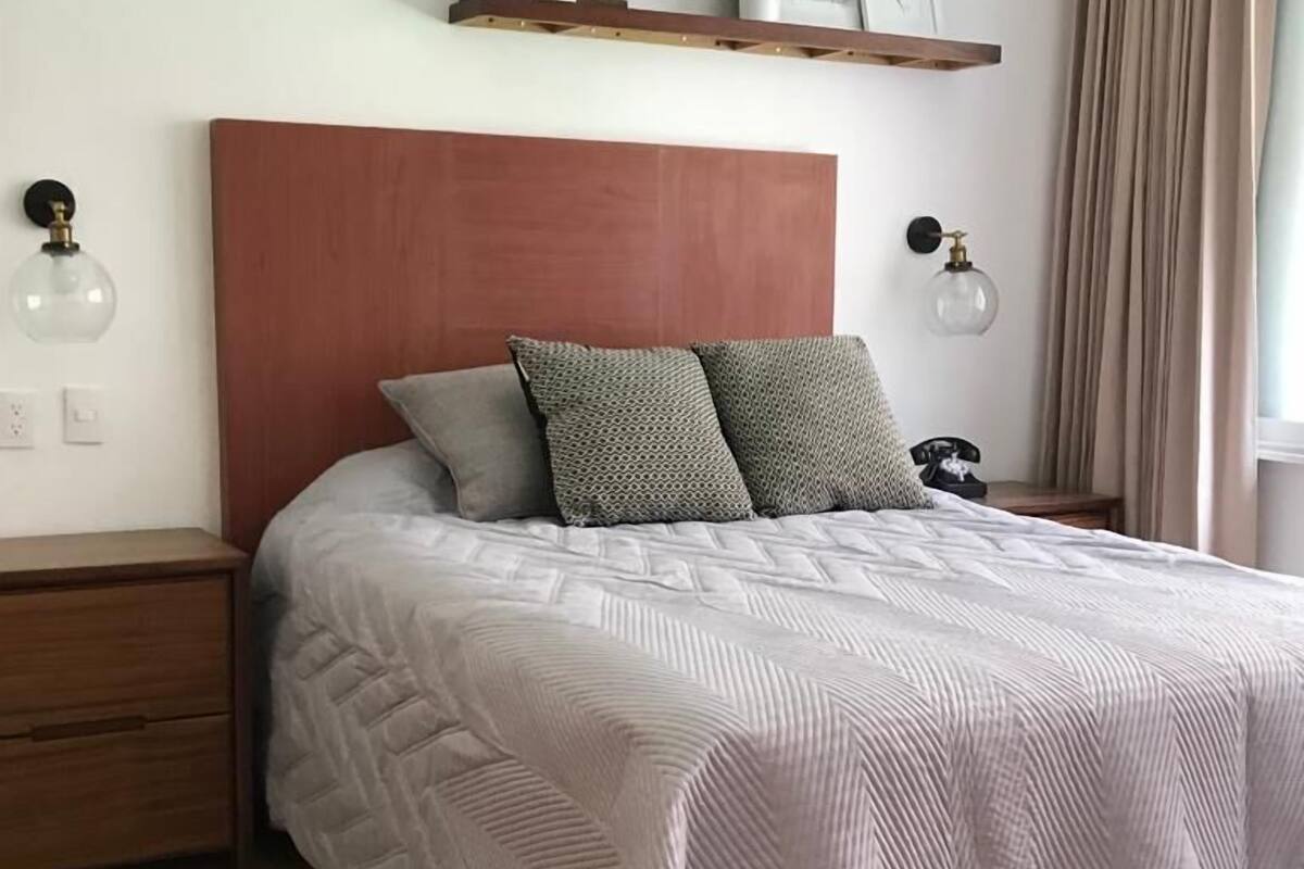 Ein Schlafzimmer mit einem hölzernen Kopfteil, einem Bett mit gemustertem Überwurf, zwei Nachttischen mit Lampen, einer Ablage mit Dekoration und einem Fenster mit Vorhängen.