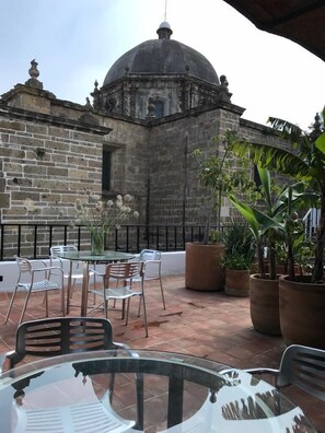 Um pátio ao ar livre com mesa e cadeiras de vidro redondas, rodeado por plantas em vasos e um edifício de pedra com cúpula ao fundo.