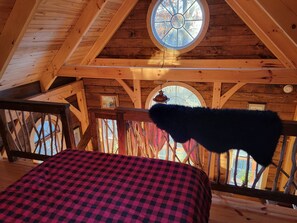 A cozy attic bedroom with a checkered bedspread, a wooden ceiling with a skylight, and a rustic wooden railing.