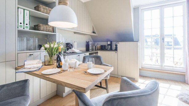 A modern kitchen with a dining area, featuring a wooden table, grey chairs, and a large window with a view. - Höög Hüs, Whg. 2A - Höög Hüs (Sylt)