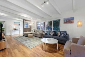 A modern living room with a sofa, a coffee table, and a rug. There are windows with a view of greenery outside.