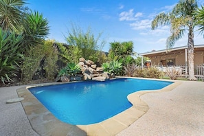 A swimming pool with a curved edge, surrounded by various plants and a rock formation.