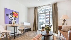 Habitació d'hotel moderna amb sòl de fusta, taula de cafè rodona amb un ram, sofà, escriptori amb cadires, llit, finestra gran amb cortines i vista a la ciutat.