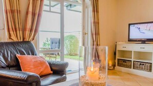 A living room with a leather sofa, a glass candle holder with a lit candle, a television, and a view of the outdoors through large windows. - Residence Bismarck, Whg. 5a - Residence Bismarck (Sylt)
