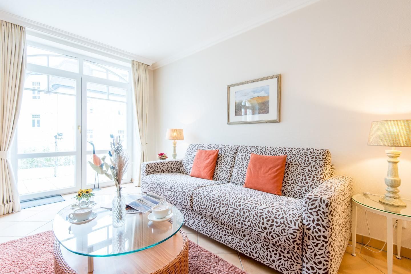 A living room with a patterned sofa, a glass coffee table, a framed picture on the wall, and a view through a large window.