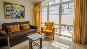 A living room with a sofa, armchair, and a glass coffee table. There is a large window with curtains and a painting on the wall. - Residence Bismarck, Whg. 12a - Residence Bismarck (Sylt)