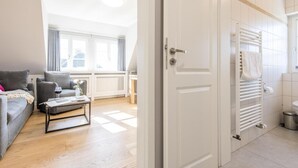 Un salon moderne avec un canapé, une table basse et une table à manger. Il y a une fenêtre avec des rideaux, un radiateur et une porte blanche qui donne sur une salle de bain équipée d’un porte-serviettes.