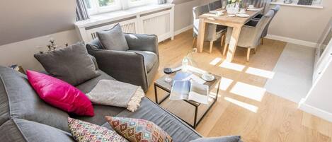 A cozy living room with a grey sofa, colorful pillows, a coffee table, and a dining area with a TV.