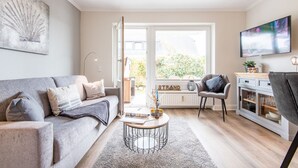 Een moderne woonkamer met een grijze bank, een bijzettafeltje, een televisie en uitzicht op het groen door een groot raam.