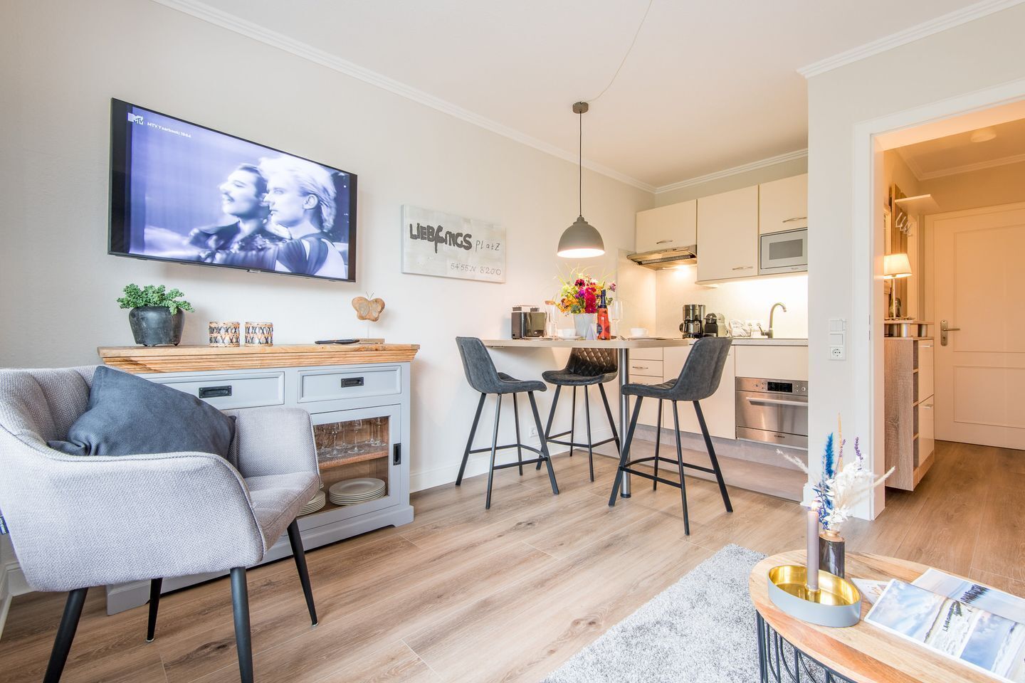 Un salon moderne avec un téléviseur, un coin repas avec des tabourets de bar et une cuisine dotée d’un plan de travail en bois.