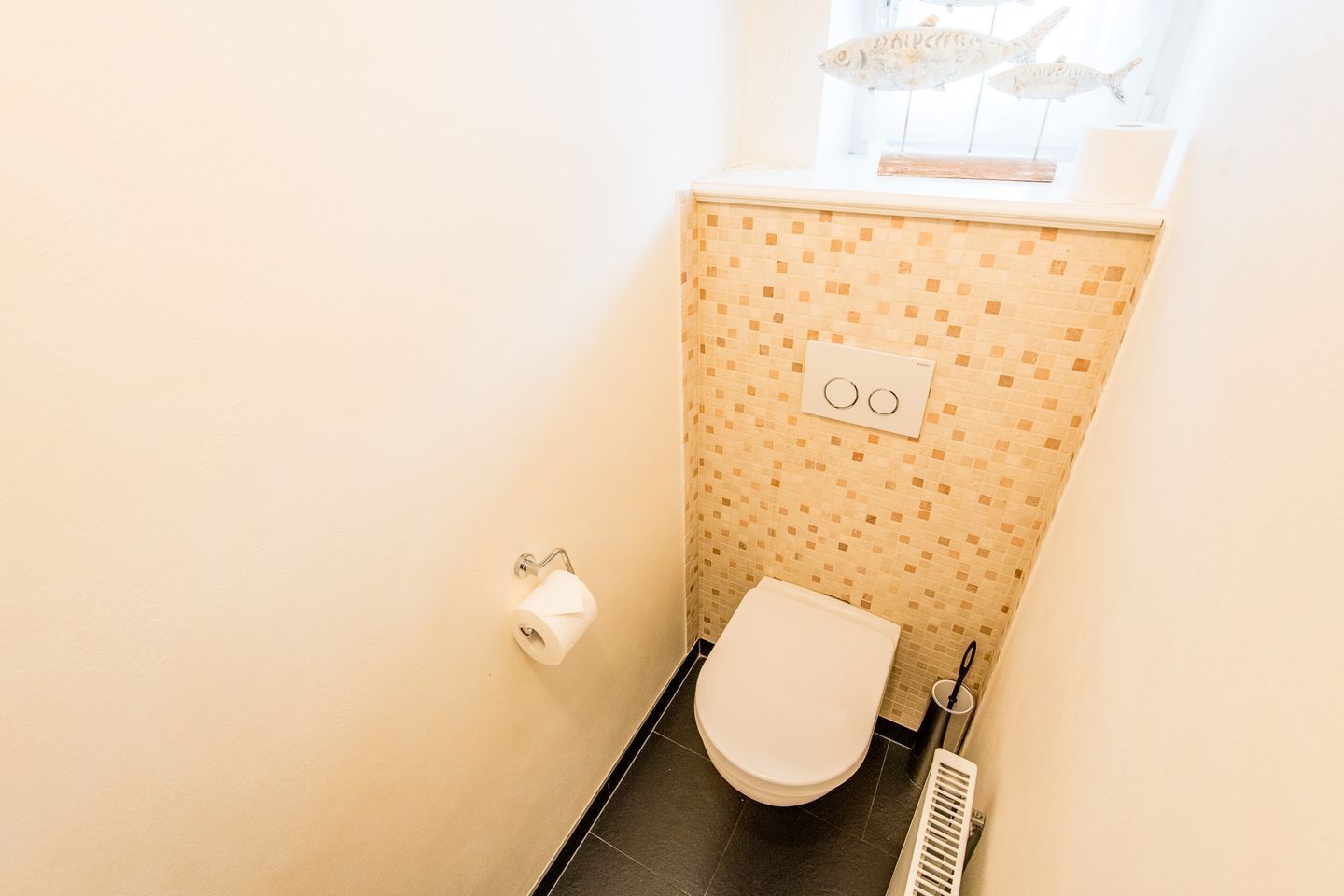 A small bathroom with a toilet, a roll of toilet paper, and a window with decorative fish.
