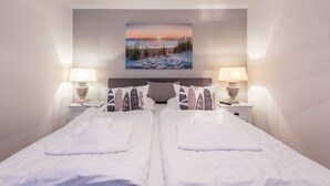 Hotelkamer met een bed, twee nachtkastjes met lampen, een schilderij van een zonsondergang boven de oceaan en een groot raam.