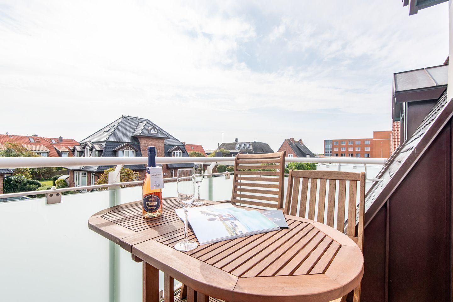 A wooden table with a bottle of wine and glasses on a balcony overlooking a residential area with houses and a bridge.