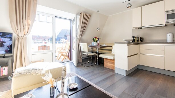 A modern hotel room with a bed, a dining area, a kitchen, and a large window with curtains. - House Nordmarkgarten, Whg. 7 - House Nordmarkgarten (Sylt)
