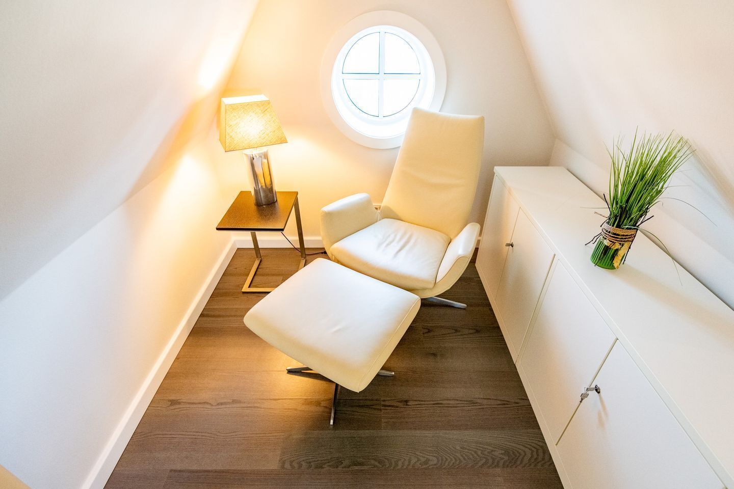 Un dormitorio moderno y luminoso en el ático, con una butaca de cuero blanco, una mesita con lámpara y una ventana con vistas.