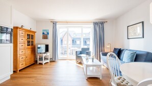 A bright living room with a wooden cabinet, a white table, a blue sofa, and a view of buildings through large windows. - House Hauke, Whg. 6 - House Hauke (Sylt)