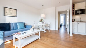 A modern living room with a blue sofa, a white coffee table, and a dining area with white chairs. A kitchen with white cabinets and a microwave is visible in the background. - House Hauke, Whg. 6 - House Hauke (Sylt)