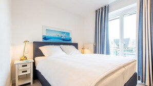 A hotel room with a bed, a nightstand with a lamp, a painting above the bed, and a window with curtains. - House Hauke, Whg. 8 - House Hauke (Sylt)