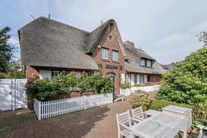 Un bâtiment traditionnel en briques, doté d’un toit de chaume, d’une clôture blanche en bois et d’un jardin planté de fleurs.