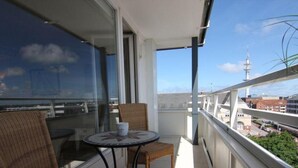 Un balcone con sedie in vimini e un tavolino rotondo, con vista su un panorama urbano che include edifici e una torre.
