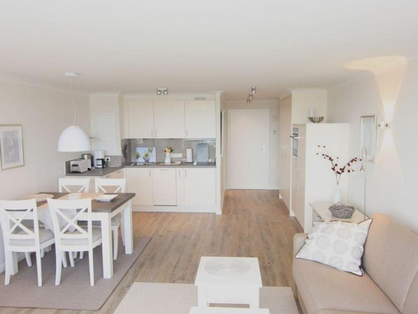 A modern kitchen with white cabinets, a dining area with white chairs, and a living room with a beige sofa and a small white table.