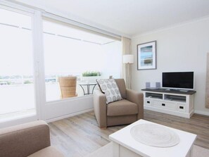 A modern living room with a sofa, armchair, and a flat-screen TV on a white stand. - Hanseatenhaus, Whg. 63 Michelsen - Hanseatenhaus (Sylt)