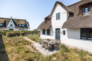 A coastal house with a thatched roof, white exterior walls, and a dining area with outdoor furniture.