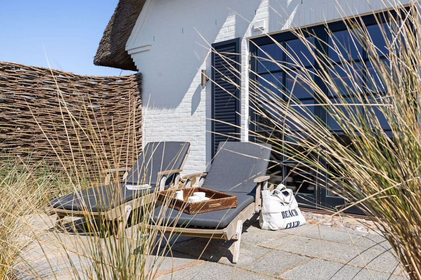 Una casa davant del mar amb teulada de palla, una cadira de relax, una taula petita i una bossa de platja.