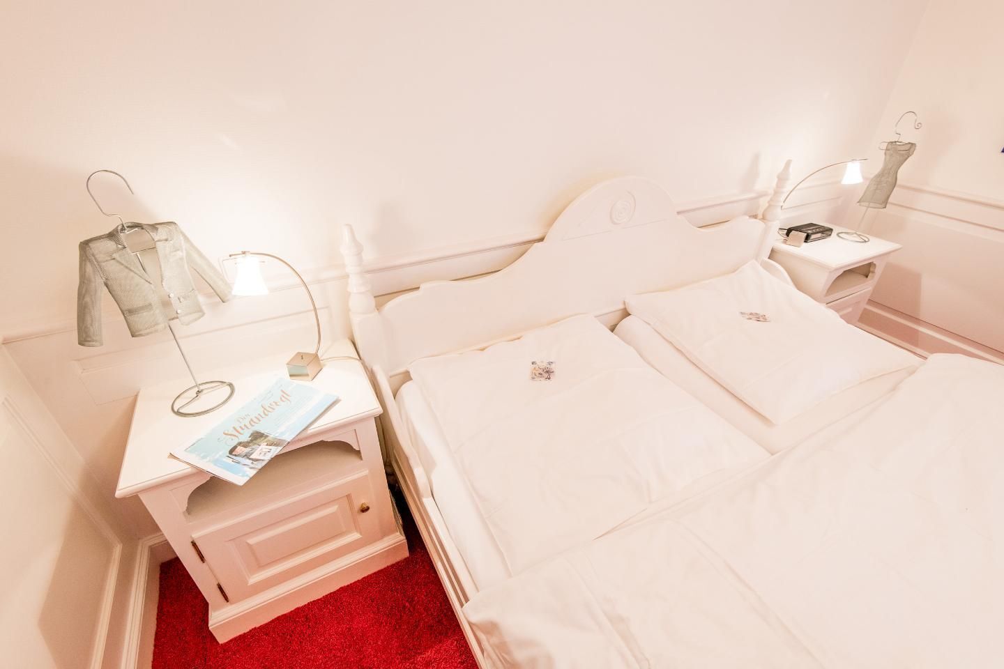A neatly made bed with white linens, a small bedside table with a magazine, and a hanging garment on a hook.