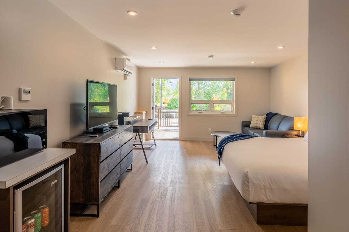 A modern hotel room with a kitchenette, a bed, a desk, and a large window offering a view of the outdoors.