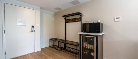 A room with a white door, a small table, a microwave on a cabinet, and a thermostat on the wall.