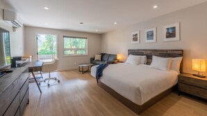 A modern hotel room with a large bed, a desk, a chair, and a balcony with a view of greenery. - Rooms at Riveredge (Golden)