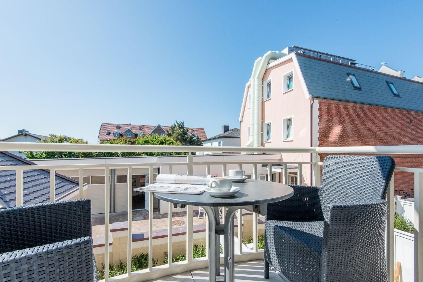 Ein Balkon mit Korbmöbeln, einem Tisch mit Tassen und Untertassen und Blick auf benachbarte Gebäude.