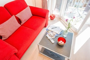 A red sofa with patterned pillows, a glass coffee table with magazines, a remote control, and a vase with flowers. - House Mateika, Whg. 10 Sammy - House Mateika (Sylt)