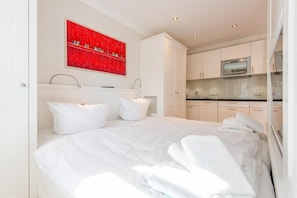 A modern hotel room with a large bed, white linens, a red artwork above the bed, and a kitchenette in the background. - House Mateika, Whg. 10 Sammy - House Mateika (Sylt)
