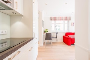 A modern kitchen with white cabinets, a granite countertop, and a built-in oven. There is a dining area with a red sofa and a table, and a window with a striped valance. - House Mateika, Whg. 10 Sammy - House Mateika (Sylt)