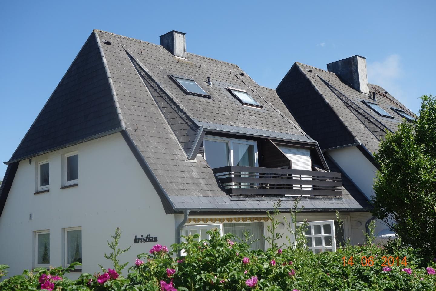 Une maison à deux étages avec un toit en pente, un balcon au deuxième étage et un jardin orné de fleurs roses à l’avant.