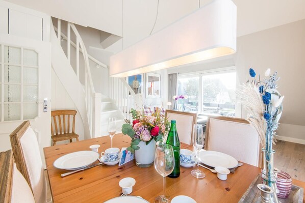 A dining area with a wooden table set for four, surrounded by white chairs. A staircase is visible in the background, leading to an upper level.