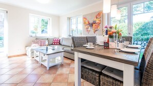Un salon moderne avec un canapé, une table à manger dressée pour deux personnes et une grande fenêtre donnant sur un paysage verdoyant.