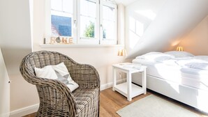 Une chambre à coucher avec un lit, une chaise en osier, une petite table de chevet blanche et une fenêtre donnant sur l’extérieur.