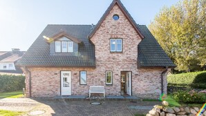 Une maison à deux étages, construite en briques, avec un toit à pignon, une porte d’entrée et un petit porche agrémenté d’un banc.