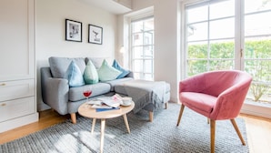 Un salon moderne avec un canapé gris, un fauteuil rose, une table basse et une grande fenêtre donnant sur un paysage verdoyant.
