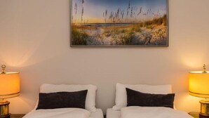 A double bed with white bedding and two black pillows, flanked by two bedside lamps. A landscape painting of a beach scene hangs above the bed. - Villa Maybach, Whg. 3 - Villa Maybach (Sylt)