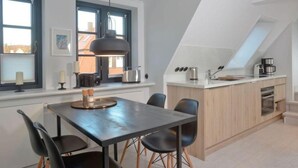 A modern kitchen with a dining area, featuring a large island, pendant lighting, and a view of the city through the windows. - Villa Maybach, Whg. 8 - Villa Maybach (Sylt)