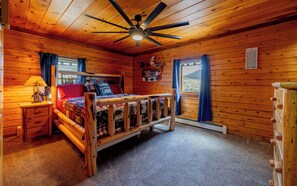 A cabin-style bedroom with a wooden ceiling, a bed with a quilt, a nightstand, a lamp, and a window with curtains.