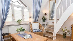 Een knus interieur met een houten tafel, een rieten stoel, een klein bijzettafeltje met een lamp en een trap.